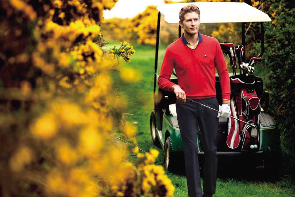 一位气质优雅的男士身着卡其色针织衫，伫立于秋日球场，手持球杆眺望远方，袖口的细腻纹路在阳光下泛着柔和光泽