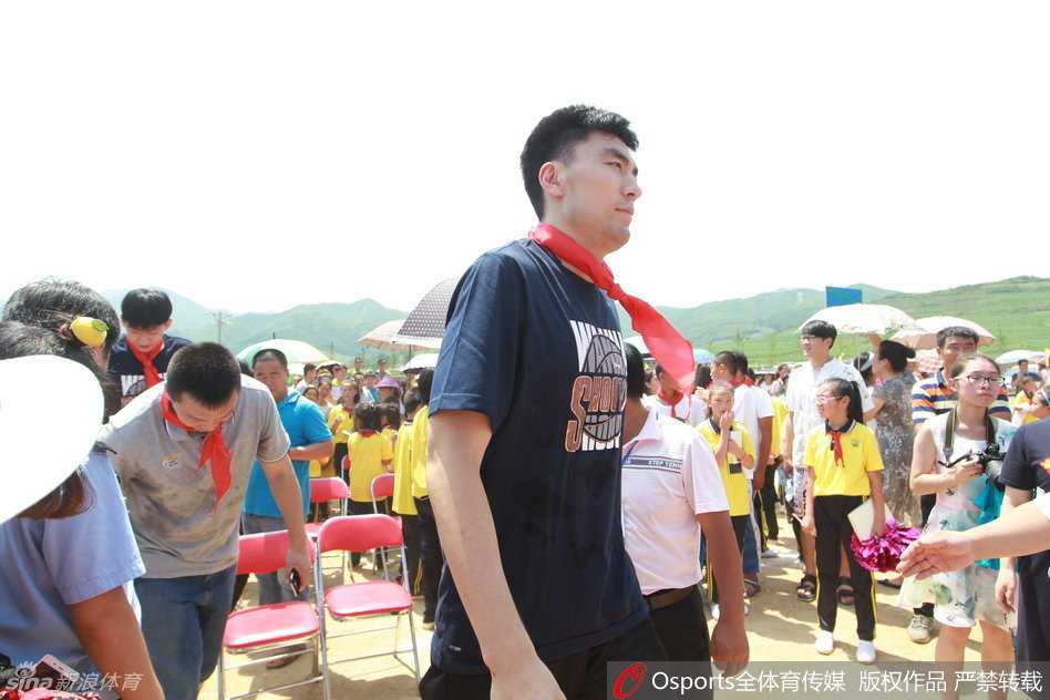 一名少先队员正在为李晓旭佩戴红领巾，李晓旭微微低头配合，脸上洋溢着温暖的笑容，旁边站满了鼓掌的师生。